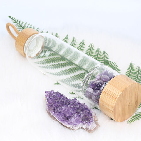 Glass and bamboo water bottle with amethyst crystals laying on a fern leaf.
